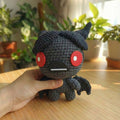 Amigurumi fatto a mano di Azazel dal videogioco The Binding of Isaac, realizzato all'uncinetto in grigio scuro e nero con ali da pipistrello e grandi occhi rossi circolari.