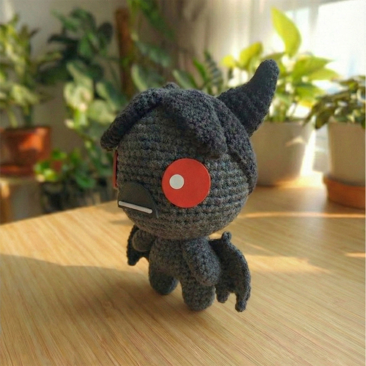 Amigurumi fatto a mano di Azazel dal videogioco The Binding of Isaac, realizzato all'uncinetto in grigio scuro e nero con ali da pipistrello e grandi occhi rossi circolari.
