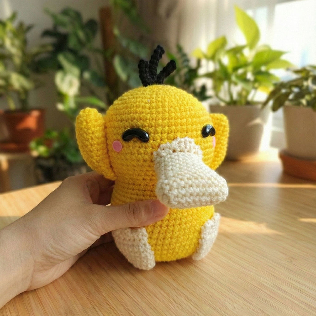 Peluche artigianale di Psyduck (Pokémon) realizzato a crochet, mostrato tenuto in mano su un tavolo di legno con piante sullo sfondo. Dettagli curati con guance rosa e occhi neri lucidi.