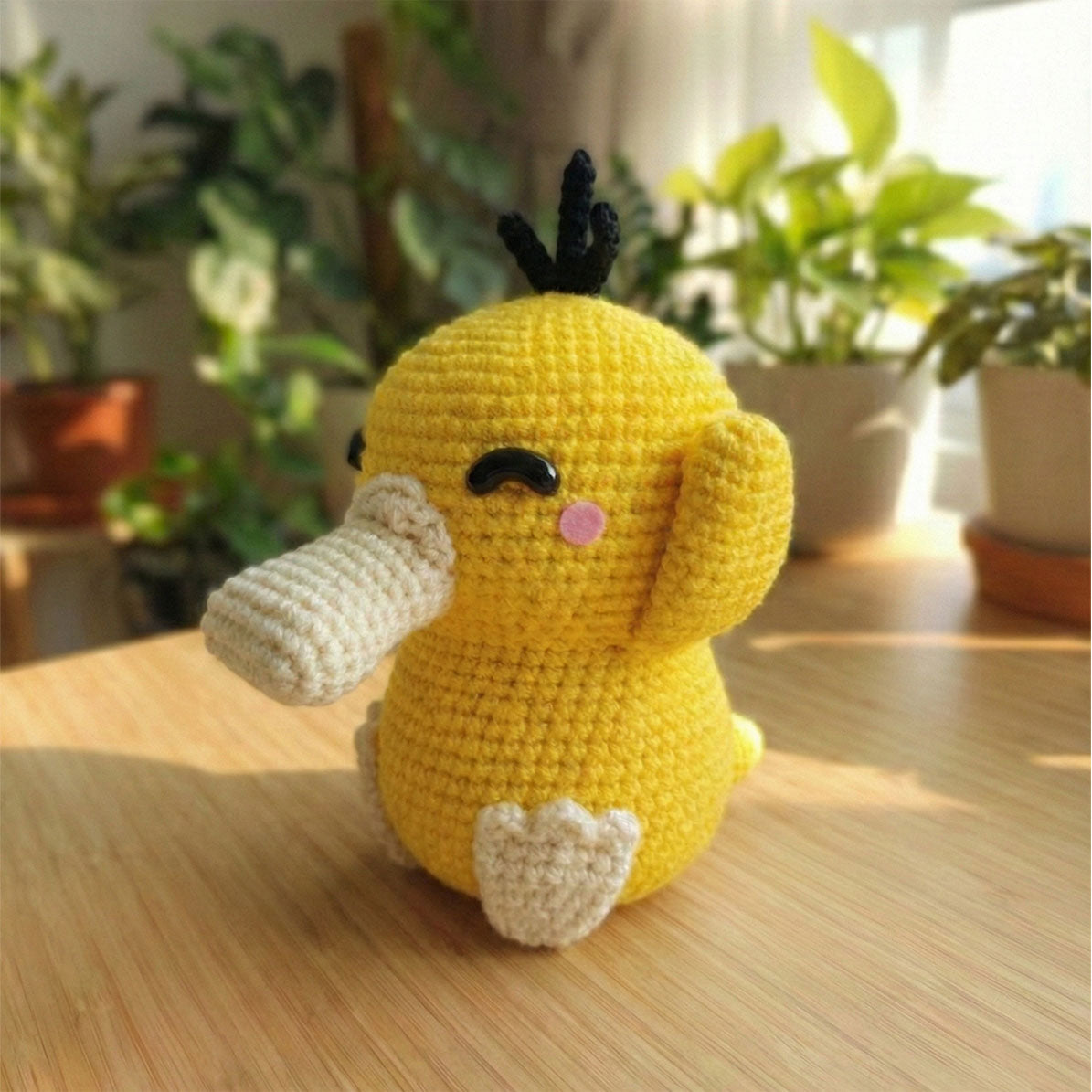 Amigurumi di Psyduck fatto a mano all'uncinetto, colore giallo brillante con becco color panna, tre ciuffetti neri sulla testa e un'espressione simpatica con un occhio ammiccante.